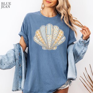 May include: A blue denim-colored t-shirt featuring a large, central graphic of a seashell composed of various patchwork patterns in yellow, white, and blue, including stripes, gingham, and floral prints. The text BLUE JEAN appears in the top left corner.
