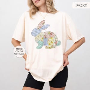 May include: An ivory t-shirt featuring a large rabbit graphic composed of various patchwork patterns, including blue gingham, yellow floral, and lace textures. The text IVORY appears in the top right corner, and MORE COLOR OPTIONS is on the left.