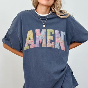 May include: A dark blue short-sleeve t-shirt featuring the word AMEN printed in a large, arched, collegiate-style font. Each letter is filled with a different pastel-colored patchwork pattern, including floral, gingham, and plaid designs.