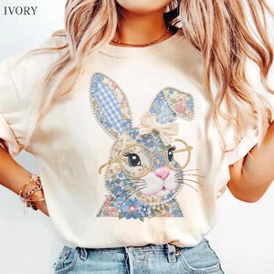 May include: An ivory t-shirt featuring a graphic of a rabbit with floral and blue gingham patterns. The rabbit wears gold-rimmed glasses and a cream-colored bow between its ears. The design is accented with a string of white pearls.