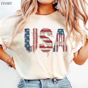 May include: An ivory short-sleeve t-shirt featuring the letters USA printed in a distressed, frayed-edge style. The letters are filled with a graphic pattern of the American flag, displaying red and white stripes and a blue field with white stars.