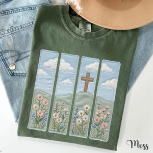 May include: An olive green crewneck t-shirt featuring four vertical rectangular panels with embroidered designs. The panels depict a light blue sky with white clouds above green hills filled with colorful wildflowers and a brown cross.