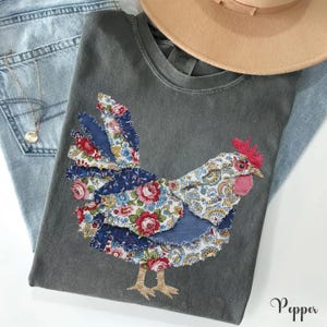 May include: A charcoal gray short-sleeve t-shirt featuring a large rooster graphic composed of various fabric patches with floral, paisley, and solid blue patterns. The rooster has a red comb and wattle. A tan hat and blue denim jeans are visible.