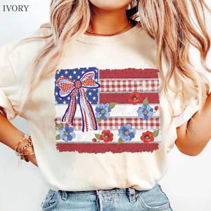 May include: An ivory short-sleeve t-shirt featuring a graphic print of a stylized American flag with a red and white gingham bow, blue and red flowers, and silver glitter accents. The design is centered on the chest of the crew neck shirt.