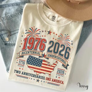 a white t-shirt with a graphic design celebrating the 1976 and 2026 Centennial Years of the United States. The design includes a map of the United States, fireworks, and the text "1976 2026" indicating the years. The t-shirt is placed on a white surface, with a pair of jeans and a hat visible in the background.