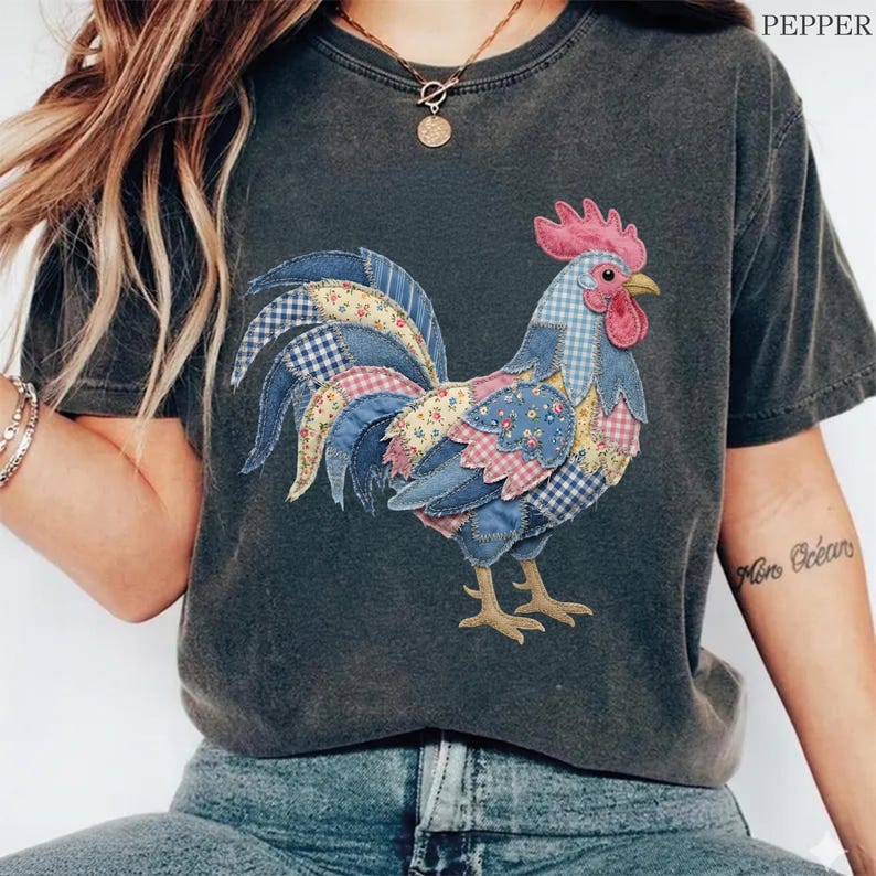 K&ouml;nnte beinhalten: Dunkelgraues T-Shirt mit einem bunten Patchwork-Hahn-Design. Der Hahn hat einen rosa Kamm, einen roten Kehllappen und einen Schwanz mit blau, gelb und rosa gemusterten Stoffen. Das Shirt hat einen Vintage-Stil, ideal f&uuml;r Freizeitkleidung.