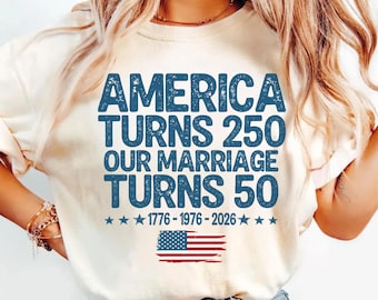 May include: An ivory t-shirt featuring blue distressed text that reads AMERICA TURNS 250 OUR MARRIAGE TURNS 50, with the dates 1776-1976-2026 and a graphic of the American flag below the text. The shirt has a crew neckline and short sleeves.