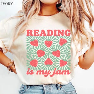 May include: An ivory t-shirt featuring a graphic print with the text READING is my jam in pink letters. The design includes a repeating pattern of red strawberries set against a background of light green radiating sunburst lines and leaves.