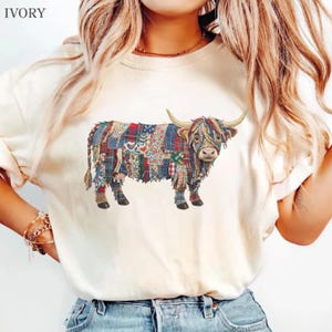 May include: An ivory t-shirt featuring a graphic of a highland cow composed of various colorful patchwork fabric patterns including plaid, floral, and denim textures. The cow is depicted in profile facing right with long horns and a shaggy coat.