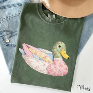 May include: Green t-shirt featuring a patchwork duck design. The duck has a checkered head, a yellow beak, and wings with floral and polka dot patterns. The shirt is paired with blue jeans and a necklace.