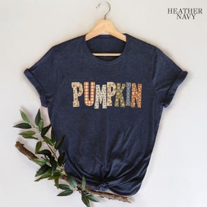 May include: A heather navy short-sleeve t-shirt featuring the word PUMPKIN printed across the chest. Each letter is filled with a different pattern, including floral, plaid, and polka dot designs in shades of orange, white, green, and blue.