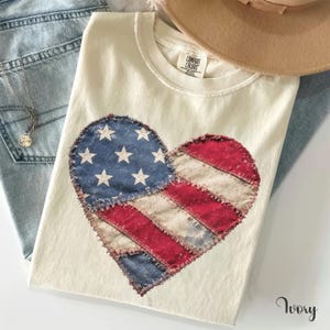 May include: An ivory t-shirt featuring a heart-shaped graphic with a distressed American flag pattern. The heart is composed of blue fabric with white stars and red and white stripes with frayed edges. The shirt has a Comfort Colors brand label.