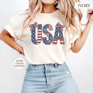 May include: An ivory t-shirt featuring the text USA in a distressed, patriotic pattern of red and white stripes, blue stars, and leopard print accents. The letters U and A are decorated with red and white striped bows at the top corners.