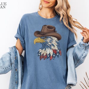May include: A blue jean colored t-shirt featuring a graphic of a bald eagle wearing a brown cowboy hat. The eagle is illustrated with a patchwork pattern of white stars, red and white plaid, and floral designs. The text BLUE JEAN is in the corner.
