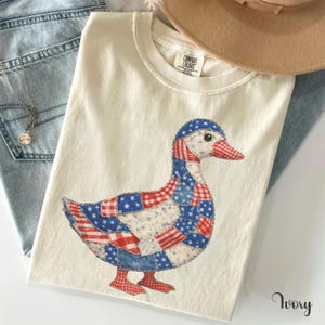May include: An ivory t-shirt featuring a graphic of a duck composed of various red, white, and blue patchwork patterns including stars and stripes. The shirt has a crew neckline and a Comfort Colors brand label visible at the inner collar area.