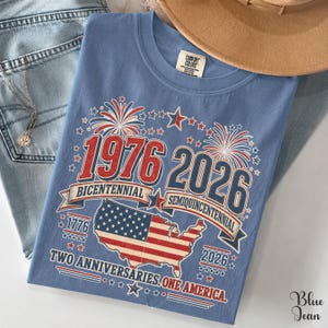 A blue t-shirt with a graphic design celebrating the 1976 and 2026 Centennial Years of the United States, featuring fireworks, stars, and the American flag.