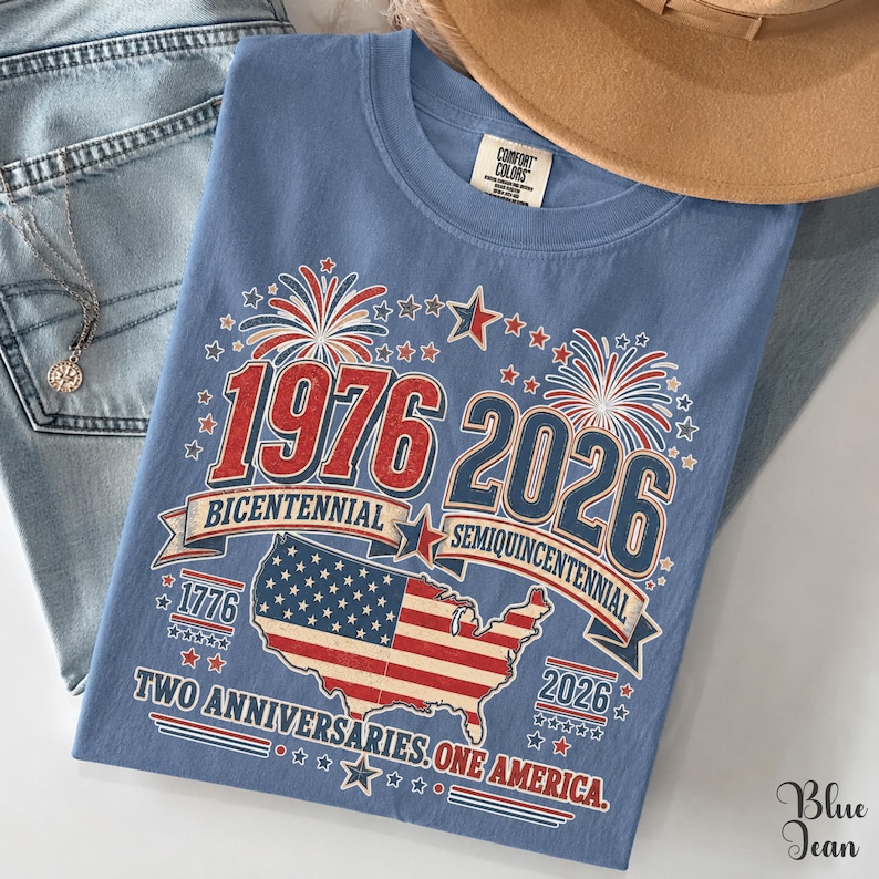 A blue t-shirt with a graphic design celebrating the 1976 and 2026 Centennial Years of the United States, featuring fireworks, stars, and the American flag.
