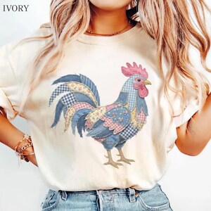 May include: An ivory t-shirt featuring a graphic of a rooster composed of various blue, pink, and yellow patchwork patterns. The rooster has a pink comb and wattle, with a light blue body and a tail made of floral, gingham, and plaid fabric designs.