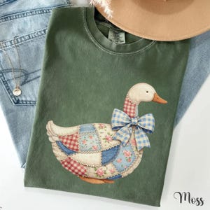 May include: A moss green crew neck t-shirt featuring a graphic of a goose with a patchwork body design. The goose is decorated with various patterns including red and white gingham, blue and white floral, and cream fabric, finished with a blue gingham bow.