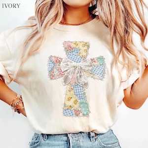 May include: An ivory short-sleeve t-shirt featuring a large cross graphic composed of various patchwork patterns including blue gingham, yellow floral, and pink rose prints. A white lace bow is centered over the middle of the cross design.