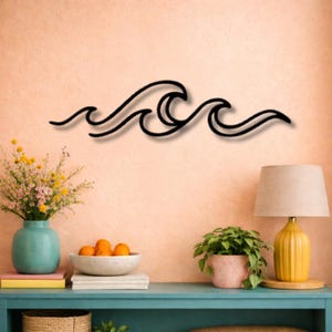 Op de afbeelding: Een wanddecoratie van zwart metaal met een doorlopende, gestileerde lijntekening van oceaangolven. Het ontwerp bestaat uit drie afzonderlijke golftoppen die met elkaar verbonden zijn door vloeiende, gebogen lijnen in een horizontaal patroon.