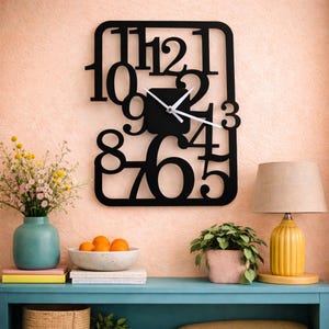 Peut inclure: Une horloge murale noire de forme carrée avec des chiffres découpés et un centre noir. Les aiguilles de l'horloge sont blanches. L'horloge est fixée sur un mur de couleur pêche. Un vase turquoise avec des fleurs, un bol d'oranges et une lampe jaune sont sur une étagère bleue en dessous.