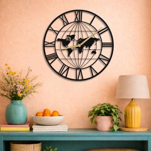 Puede incluir: Un reloj de pared circular de metal negro con una silueta de mapa mundial en el centro y grandes números romanos en el perímetro. El reloj tiene dos manecillas finas de color dorado y un patrón de cuadrícula que representa meridianos.