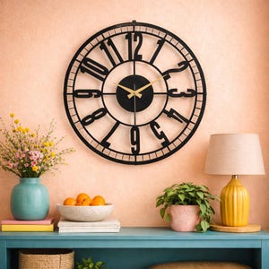Puede incluir: Un gran reloj de pared redondo de metal negro con números romanos. El reloj tiene manecillas doradas y está montado en una pared de color melocotón. El reloj es el punto focal de una habitación, con una estantería azul debajo.