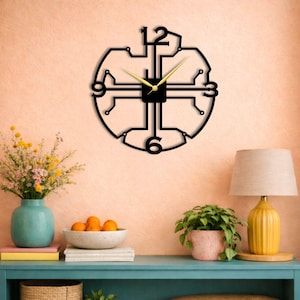 May include: Black metal wall clock with gold-colored hands and a unique circuit board design. The clock features the numbers 12, 3, 6, and 9. The clock is mounted on a peach-colored wall.