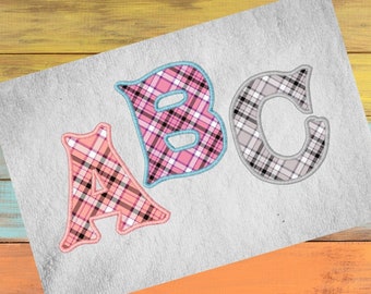 May include: Three embroidered letters, A, B, and C, in a plaid pattern with pink, black, and grey hues. The letters are on a light grey fabric, set against a backdrop of colourful wooden planks.