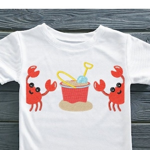 May include: White t-shirt with a red bucket, yellow shovel, and two red crabs with black eyes. The crabs are on either side of the bucket.