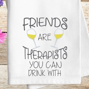 Puede incluir: Toalla de cocina blanca con el texto "Friends are therapists you can drink with" y dos copas de vino con líquido amarillo.