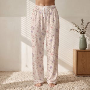 May include: Full-length lounge pants in a light cream color featuring a delicate floral pattern in shades of pink and purple. The pants have an elasticized waistband with a white drawstring tie at the center front and a loose, straight-leg fit.