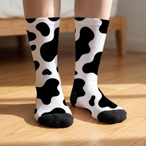 May include: A pair of crew-length socks featuring a white base with an all-over black cow print pattern. The socks have solid black toe sections and a ribbed cuff at the top, extending to the mid-calf area with a consistent spotted design.
