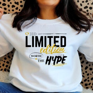 May include: White sweatshirt featuring the text "LIMITED edition WORTH THE HYPE" in black and gold. The design includes arrows and a star. The sweatshirt is long-sleeved with a crew neck.