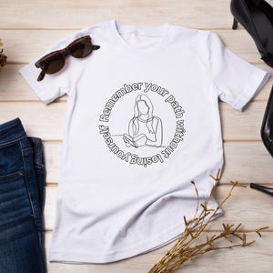 May include: White t-shirt with a circular line drawing of a person reading, with the text "Remember your path without losing yourself." Also includes blue jeans, sunglasses, and black high heels.
