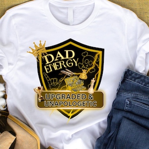 May include: A white t-shirt featuring a black and gold shield graphic with the text DAD ENERGY and UPGRADED & UNAPOLOGETIC. The design includes a crown, a skull, a money bag, a trophy, a toolbox, a chess piece, and lightning bolt accents.