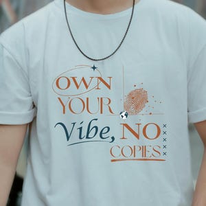 Op de afbeelding: Wit t-shirt met de tekst "OWN YOUR VIBE, NO COPIES" in oranje en blauw. Het ontwerp bevat een vingerafdruk en een wereldbol-icoon. Het shirt heeft korte mouwen en een ronde hals.