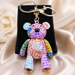 Op de afbeelding: Een goudkleurige metalen sleutelhanger in de vorm van een teddybeer, ingelegd met veelkleurige strass-steentjes. De beer heeft een roze gezicht, een hartvormige kristallen patch op de borst en ledematen in tinten blauw, groen, geel en paars.