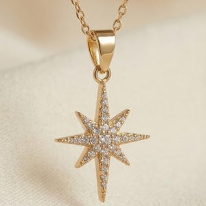May include: A gold-coloured necklace with a starburst pendant. The pendant is set with small, clear gemstones. The necklace has a delicate chain and a simple clasp. The background is a soft, off-white fabric.