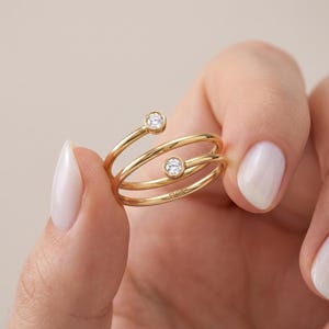 Könnte beinhalten: Ein goldener Wickelring mit zwei offenen Enden, die jeweils mit einem einzelnen runden, klaren Stein in einer Zargenfassung besetzt sind. Das Band hat eine glatte, polierte Oberfläche und das Wort FIOR ist auf der Innenseite eingraviert.