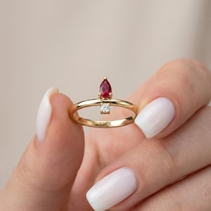 May include: A gold ring with a pear-shaped, deep red gemstone and a small, square-cut diamond. The ring has a delicate band and the word "FIOR" is engraved on the band. The ring is held by a person.
