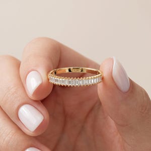 May include: A gold band ring featuring a row of clear baguette-cut stones set horizontally. The metal band has a beaded texture along the top and bottom edges of the stone setting. The interior of the ring is engraved with the text FIOR.