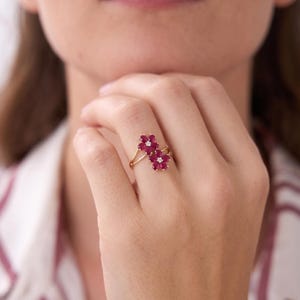 Puede incluir: Un anillo de oro con dos racimos en forma de flor, cada uno compuesto por cinco p&eacute;talos de rub&iacute; rojo intenso que rodean una peque&ntilde;a piedra central clara y brillante. La banda es fina y pulida, mostrando un dise&ntilde;o delicado en el dedo.