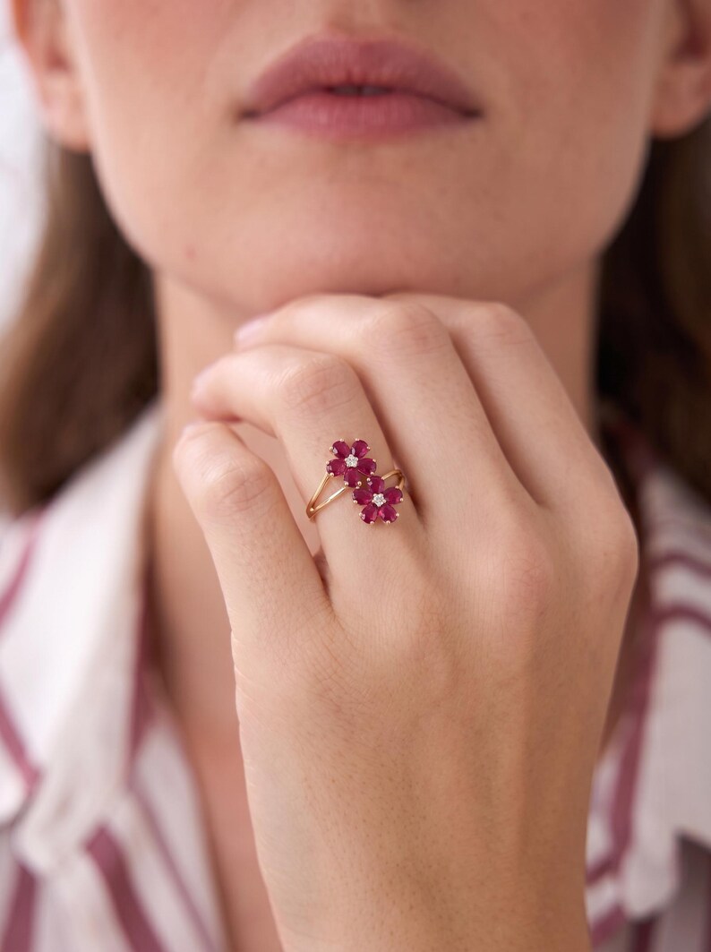 Puede incluir: Un anillo de oro con dos racimos en forma de flor, cada uno compuesto por cinco p&eacute;talos de rub&iacute; rojo intenso que rodean una peque&ntilde;a piedra central clara y brillante. La banda es fina y pulida, mostrando un dise&ntilde;o delicado en el dedo.