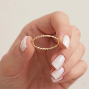 May include: A thin, gold-colored ring featuring a delicate, twisted rope-like texture. The band is circular and maintains a consistent, slender width throughout its entire circumference, showcasing a polished, metallic finish with a subtle sheen.