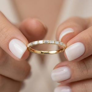 May include: A gold ring featuring a line of rectangular-cut diamonds. The ring is held between two hands with white nail polish. The word "FIOR" is engraved on the ring.