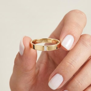 Könnte beinhalten: Ein polierter Gelbgoldring mit einem einzelnen, vertikal gefassten Diamanten im Baguetteschliff in der Mitte. Die Innenseite des Rings ist deutlich mit dem Markennamen FIOR in Großbuchstaben graviert, was auf der Metalloberfläche sichtbar ist.