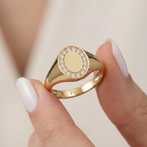 Pode incluir: Um anel de sinete dourado com uma placa central oval rodeada por um halo de pequenas pedras transparentes de corte redondo. A banda é lisa e polida, com a palavra FIOR gravada na superfície interior do aro do anel.
