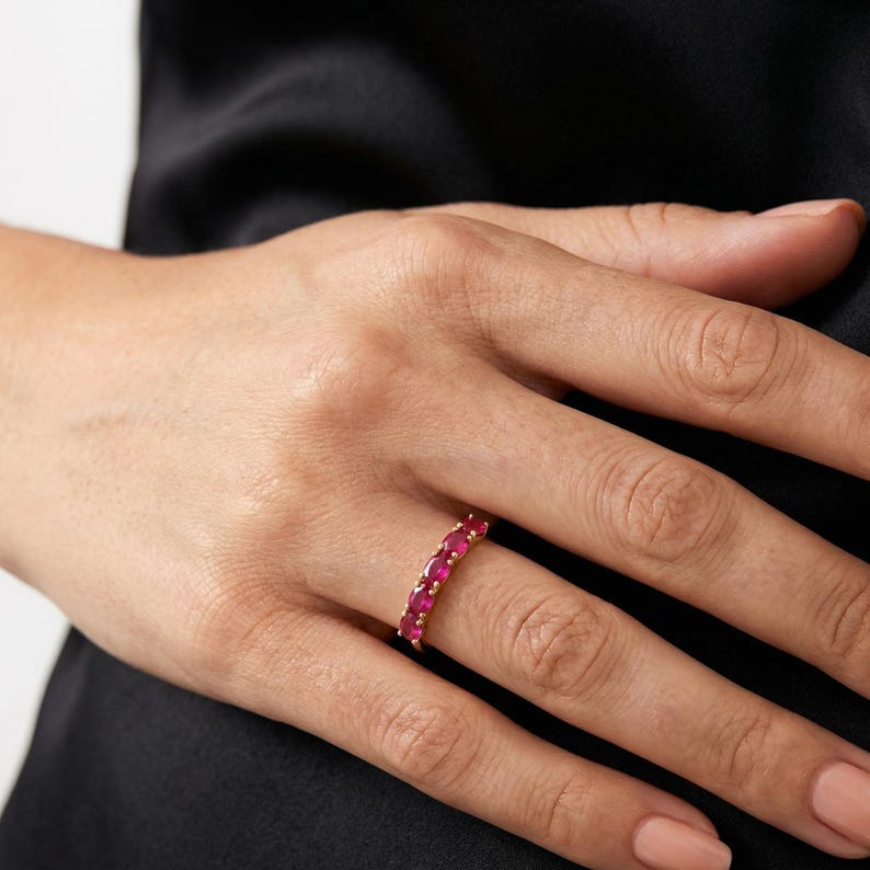 Op de afbeelding: Een gouden ring bezet met levendige rode edelstenen. De ring heeft een doorlopende rij ovaal geslepen robijnen, wat een gedurfde en elegante uitstraling cre&euml;ert. De ring wordt getoond op een hand met een zwarte achtergrond.